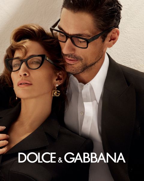 Eine Frau und ein Mann mit schicken Brillen lehnen dicht beieinander. Sie trägt DG-Ohrringe und ein schwarzes Outfit, er trägt einen schwarzen Anzug mit weißem Hemd. Unten ist das Logo von Dolce & Gabbana zu sehen.