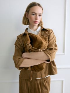 Eine Frau mit blondem Bob, die ein braunes Outfit trägt, steht mit verschränkten Armen vor einer weißen Wand und hält eine große braune Wildledertasche in der Hand.