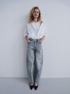 Eine Frau in einem weißen Hemd und lockeren grauen Jeans steht mit den Händen in den Taschen vor einer weißen Wand, trägt schwarze Pumps und blickt in die Kamera.