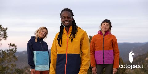 Drei Menschen in bunten Outdoor-Jacken stehen in einer Naturlandschaft mit Bäumen und Bergen. Der Himmel ist bedeckt. Unten rechts ist das Markenlogo „Cotopaxi“ zu sehen.