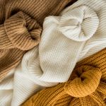 Eine Nahaufnahme von Strickpullovern in drei Farben: Braun, Creme und Senfgelb. Die Pullover sind gefaltet und lässig übereinander gestapelt, wodurch ihre strukturierten Muster und warmen Farbtöne zur Geltung kommen.