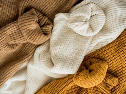 Eine Nahaufnahme von Strickpullovern in drei Farben: Braun, Creme und Senfgelb. Die Pullover sind gefaltet und lässig übereinander gestapelt, wodurch ihre strukturierten Muster und warmen Farbtöne zur Geltung kommen.