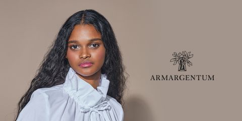 Eine Frau mit langen, welligen Haaren trägt eine weiße Bluse mit hohem Kragen. Sie steht vor einem beigen Hintergrund, auf dessen rechter Seite sich das Logo „ARMARGENTUM“ mit einem Blumenmuster befindet.