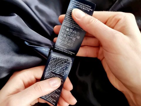 Hände halten ein dunkles Kleidungsetikett mit Pflegehinweisen, darunter Symbole zum Waschen, Bleichen, Trocknen und Bügeln. Das Etikett ist mit weißem Text auf dunklem Hintergrund versehen und das Kleidungsstück scheint aus seidigem Stoff zu bestehen.