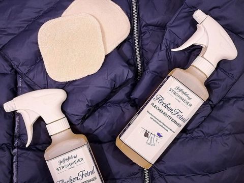 Auf einer dunkelblauen Steppjacke sind zwei Flaschen Reinigungsspray mit der Aufschrift „FleckenFeind“ platziert, daneben zwei beigefarbene Mehrweg-Reinigungspads. Die Sprühdüsen sind weiß, und die Anordnung suggeriert einen Fokus auf die Wäschepflege.