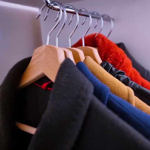 Eine Reihe Mäntel hängt auf hölzernen Kleiderbügeln in einem Schrank. Die Kleidungsstücke sind in verschiedenen Farben erhältlich, wie Schwarz, Marineblau, Senfgelb und Orange, was auf eine vielfältige Auswahl an Kleidungsstücken für verschiedene Stile oder Jahreszeiten schließen lässt.