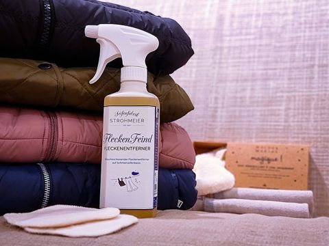Auf einem Stapel gefalteter, bunter Jacken steht eine Sprühflasche mit der Aufschrift „Flecken-Feind Fleckenentferner“. Daneben liegen Stoffmuster und eine Schachtel. Der Hintergrund weist eine strukturierte Oberfläche auf.