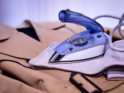 Ein blau-silbernes Bügeleisen liegt auf einer beigefarbenen Jacke, die auf einer flachen Oberfläche liegt. Ein aufgerolltes Kabel geht vom Bügeleisen aus. Die Jacke ist mit einer Gürtelschlaufe und einer Schnalle versehen.