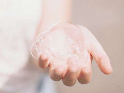 Die Hand einer Person hält eine Ansammlung zarter, durchscheinender Seifenblasen vor einem weichen, unscharfen Hintergrund. Die Blasen erscheinen schillernd mit Andeutungen von Lichtreflexionen.