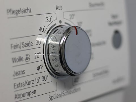 Nahaufnahme eines Waschmaschinenwählrads mit verschiedenen Einstellungen, darunter „Fein/Seide“, „Wolle“, „Jeans“ und „Extra Kurz 15' 30°“. Das Wählrad ist in der Nähe von „Wolle“ auf 30° eingestellt. Deutscher Text beschriftet verschiedene Waschprogramme und Temperaturen.