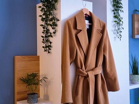 An einer weißen Wand hängt ein brauner Mantel an einem Haken, flankiert von kleinen Grünpflanzen. Die Wand und die Umgebung sind mit noch mehr Grün dekoriert, was eine frische, natürliche Atmosphäre schafft.