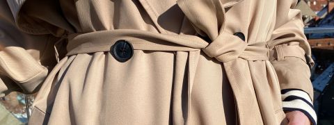 Nahaufnahme einer Person, die einen beigen Trenchcoat mit zwei sichtbaren schwarzen Knöpfen und einem gebundenen Gürtel trägt. Die Hand der Person ist seitlich teilweise sichtbar. Im Hintergrund sind verschwommene Andeutungen von Dächern zu sehen.