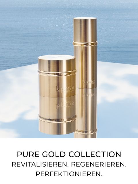 Zwei goldene, zylindrische Kosmetikbehälter aus der „Pure Gold Collection“ stehen auf einer reflektierenden Oberfläche vor einem ruhigen Wasserhintergrund. Der Text lautet: „PURE GOLD COLLECTION REVITALISIEREN. REGENERIEREN. PERFEKTIONIEREN.“.