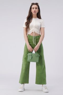 Eine Person mit langen Haaren steht vor einem schlichten Hintergrund und trägt ein weißes, gerüschtes bauchfreies Oberteil und grüne Hosen mit weitem Bein. Sie hält eine kleine grüne Handtasche und trägt weiße Turnschuhe. Auf ihrem Kopf sitzt eine Sonnenbrille.