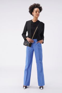 Eine Frau steht selbstbewusst vor einem schlichten Hintergrund. Sie trägt einen schwarzen Blazer, blaue Hosen und schwarz-weiße Schuhe. Sie hält eine schwarze Umhängetasche und hat eine Afrofrisur.