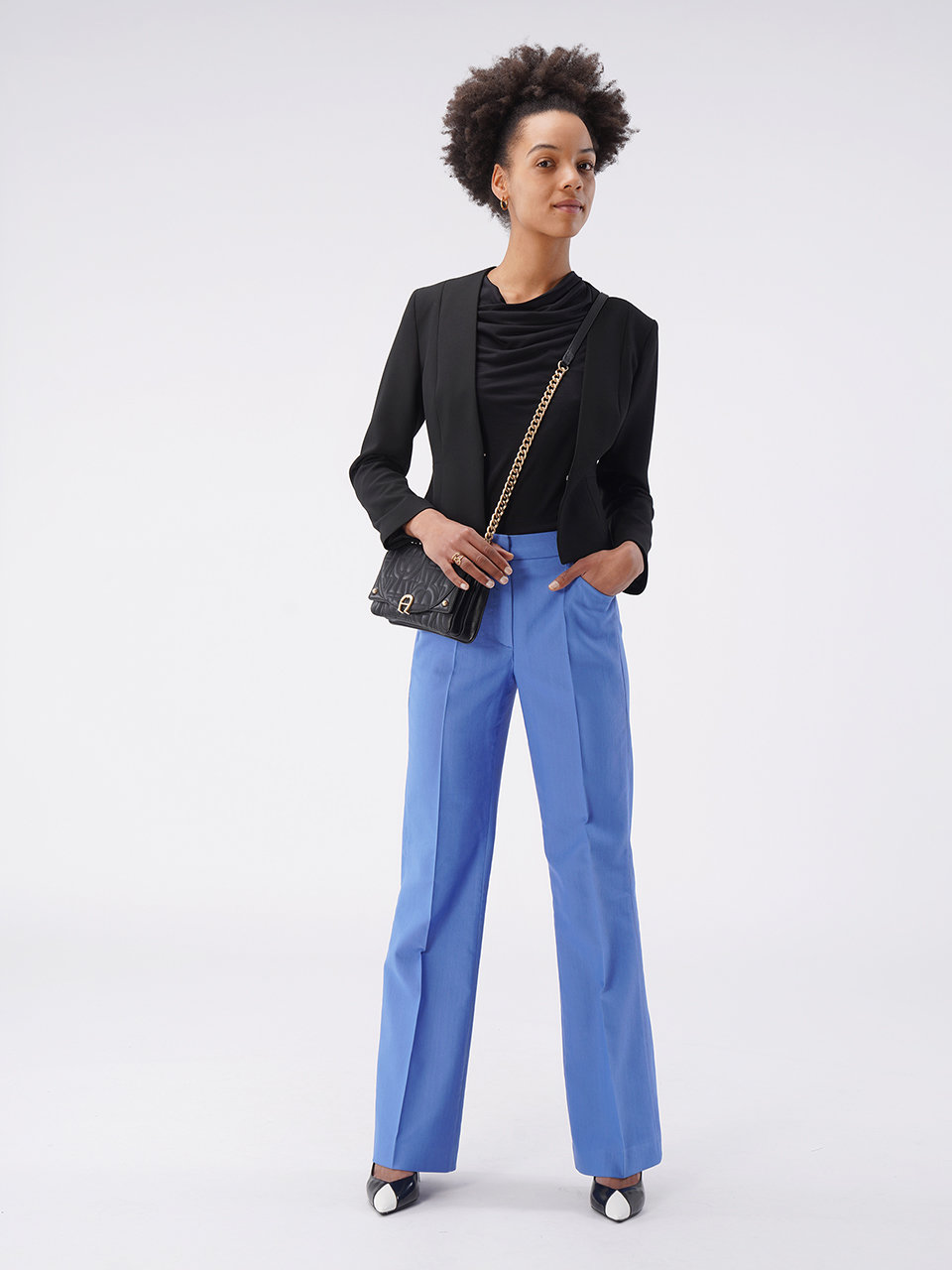 Eine Frau steht selbstbewusst vor einem schlichten Hintergrund. Sie trägt einen schwarzen Blazer, blaue Hosen und schwarz-weiße Schuhe. Sie hält eine schwarze Umhängetasche und hat eine Afrofrisur.