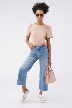 Eine Person mit lockigem Haar steht selbstbewusst mit Sonnenbrille da, trägt ein hellrosa Oberteil und blaue Jeans mit weitem Bein. Sie hält eine passende rosa Handtasche in der Hand und trägt weiße Turnschuhe mit beigen Details. Der Hintergrund ist schlicht weiß.