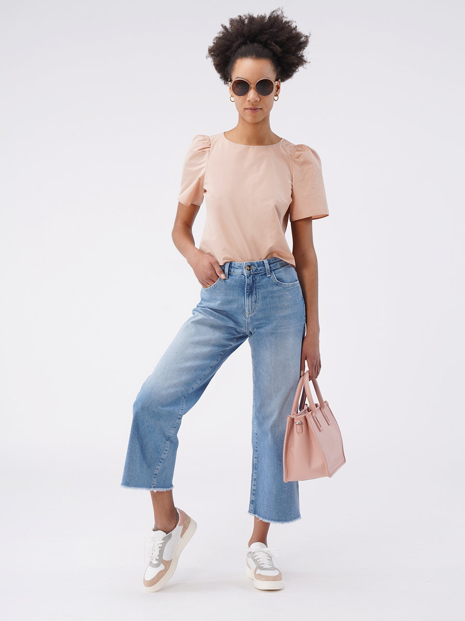Eine Person mit lockigem Haar steht selbstbewusst mit Sonnenbrille da, trägt ein hellrosa Oberteil und blaue Jeans mit weitem Bein. Sie hält eine passende rosa Handtasche in der Hand und trägt weiße Turnschuhe mit beigen Details. Der Hintergrund ist schlicht weiß.