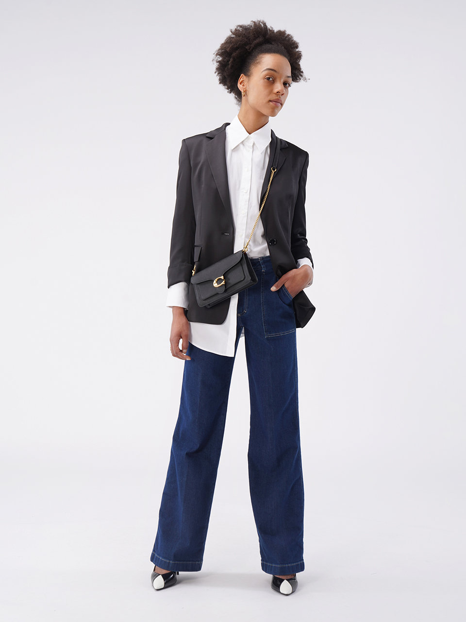 Eine Person mit kurzen lockigen Haaren trägt einen schwarzen Blazer über einem weißen Hemd, weite Bluejeans und schwarz-weiße Schuhe. Sie hält eine schwarze Umhängetasche und steht vor einem schlichten weißen Hintergrund.