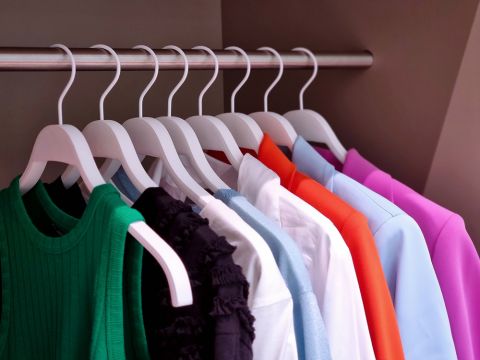 Eine Reihe bunter Kleidungsstücke auf weißen Kleiderbügeln ist ordentlich auf einer Metallstange angeordnet. Die Kleidungsstücke sind in verschiedenen Farben wie Grün, Schwarz, Weiß, Grau, Blau, Orange und Rosa erhältlich und sorgen so für eine lebendige und ordentliche Präsentation.