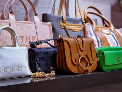 Eine Ausstellung verschiedener Designerhandtaschen, darunter eine weiße Tasche von Calvin Klein, eine braune Ledertasche mit Kettenriemen, eine grüne Clutch und mehrere Tragetaschen in unterschiedlichen Farben und Mustern, auf einem Regal angeordnet.