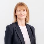 Frau mit schulterlangem blondem Haar, die einen dunklen Blazer über einem weißen Oberteil trägt, steht vor einem schlichten hellen Hintergrund. Sie blickt mit neutralem Gesichtsausdruck in die Kamera.