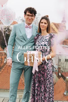 Ein Mann im hellblauen Anzug und eine Frau im geblümten Kleid stehen zusammen auf einem Dach. Sie halten ein mit einer Schleife verpacktes Geschenk in den Händen. Im Hintergrund sind Dächer und ein Turm zu sehen. Links sind rosa Luftballons zu sehen. Der Text auf dem Bild lautet: „Alles für die Gäste.“.