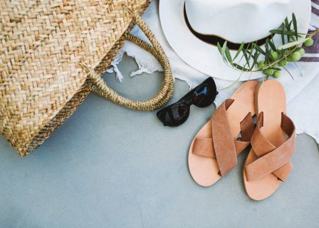 Strandutensilien wie eine Tragetasche aus geflochtenem Stroh, eine schwarze Sonnenbrille, ein weißer Sonnenhut mit Olivenzweigen und ein Paar Ledersandalen sind auf einer hellen Oberfläche angeordnet. Ein Fransentuch bedeckt teilweise den Griff der Tragetasche.