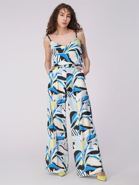 Eine Person mit welligem Haar trägt einen bunten, gemusterten Overall mit abstrakten Mustern in Blau, Schwarz und Weiß. Sie hat die Hände in den Taschen und steht vor einem schlichten Hintergrund. Sie trägt gelbe Schuhe.