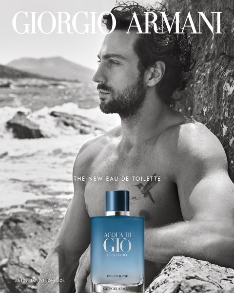 Ein Mann mit nacktem Oberkörper und lockigem Haar sitzt nachdenklich am felsigen Meeresufer. Im Vordergrund steht eine Flasche Giorgio Armani Acqua di Giò Profondo Eau de Toilette. Das Bild zeigt das Giorgio Armani-Logo.