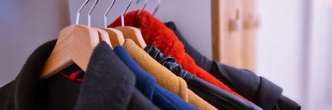 Eine Nahaufnahme mehrerer Mäntel, die auf hölzernen Kleiderbügeln in einem Schrank hängen. Die Mäntel sind in verschiedenen Farben und Materialien gehalten, darunter Schwarz, Rot, Blau und Braun, was auf eine Wintergarderobe schließen lässt. Der Hintergrund zeigt eine Teilansicht des hölzernen Schranks.
