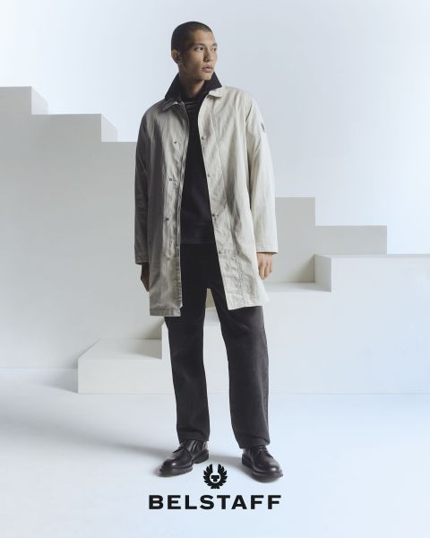 Eine Person, die einen hellen Mantel über dunkler Hose und Schuhen trägt, steht vor einem minimalistischen, geometrischen Hintergrund. Am unteren Bildrand ist das Logo „Belstaff“ zu sehen.