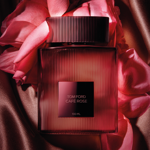 Eine Flasche des Parfüms Tom Ford Café Rose ist von rosa Rosenblättern auf seidigem, bronzefarbenem Stoff umgeben. Die Flasche ist rechteckig mit vertikalen Rillen und einem dunklen, quadratischen Etikett in der Mitte, das Luxus und Eleganz vermittelt.