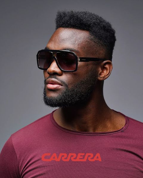 Mann trägt große quadratische Sonnenbrille und ein weinrotes Hemd mit „CARRERA“-Logo und blickt zur Seite, vor einem grauen Hintergrund.