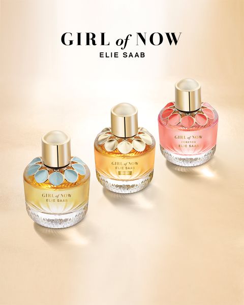 Drei Parfümflakons „Girl of Now“ von Elie Saab werden auf einer hellgoldenen Oberfläche präsentiert, jeder mit einem einzigartigen Farbschema: blaue, goldene und rosa Akzente. Die Verschlüsse sind goldfarben und der Markenname mit dem Kollektionstitel steht über den Flakons.