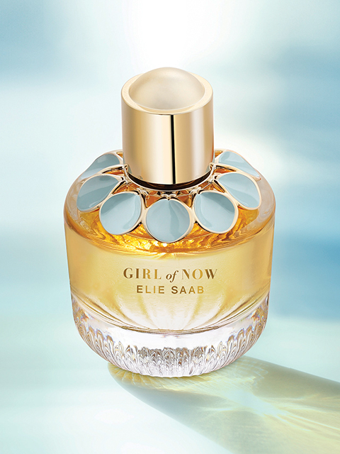 Eine Glasflasche mit „Girl of Now“ von Elie Saab steht vor einem weichen, ätherischen Hintergrund. Die Flasche weist goldene Akzente und ein Blumenmotiv um den Hals auf. Der Duft enthält eine warme, bernsteinfarbene Flüssigkeit, die für ein luxuriöses und elegantes Aussehen sorgt.