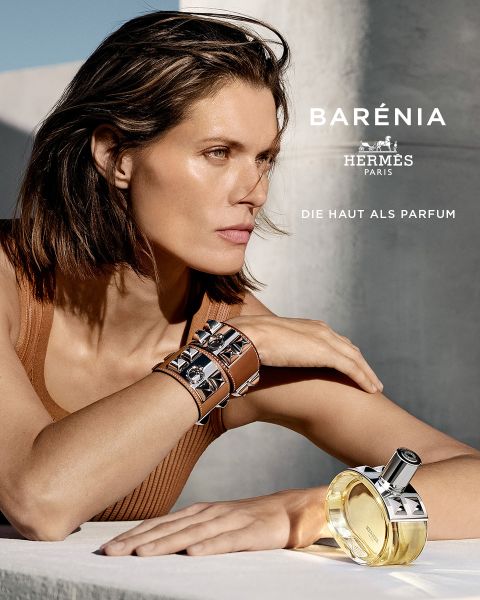 Eine Person mit schulterlangem Haar lehnt auf einer Oberfläche, trägt ein ärmelloses Oberteil und ein Armband mit metallischen Elementen. Im Vordergrund steht eine Flasche Barénia-Parfüm von Hermès Paris. Der Hintergrund ist sanft beleuchtet und es ist Text vorhanden.