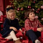 Ein kleines Mädchen und ein kleiner Junge sitzen auf dem Boden vor einem Weihnachtsbaum und lächeln, während sie rote Geschenkpakete auspacken.