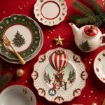 Geschirr mit Weihnachtsmotiven, darunter Teller mit einem Weihnachtsbaum und einem Heißluftballon, eine dazu passende Teekanne und eine goldene Gabel auf einer roten Tischdecke.