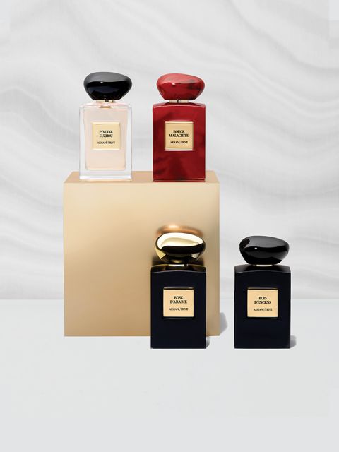 Vier elegante Parfümflakons, die auf einem goldenen Sockel ausgestellt sind. Die Flakons sind schwarz, rot und golden und haben jeweils einen passenden Verschluss. Sie sind vor einem weichen, welligen grau-weißen Hintergrund angeordnet.