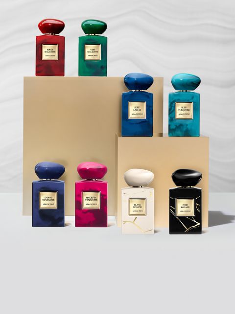 Acht Parfümflakons in verschiedenen Farben, darunter Rot, Grün, Blau, Rosa und Schwarz, sind auf beigen Podesten vor einem hellen Hintergrund angeordnet. Jeder Flakon trägt ein Etikett mit goldener Schrift.