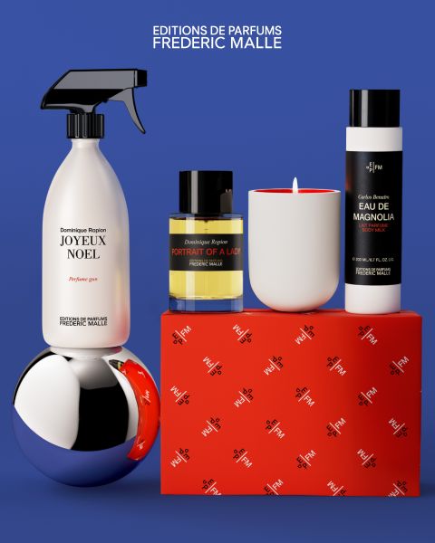Ein Display von Frederic Malle Parfüm Produkte, einschließlich einer Sprühflasche, Parfüm-Flasche, Kerze und Lotionen, auf einem roten Kasten mit einem glänzenden silbernen Ornament angeordnet, die alle vor einem blauen Hintergrund gesetzt.