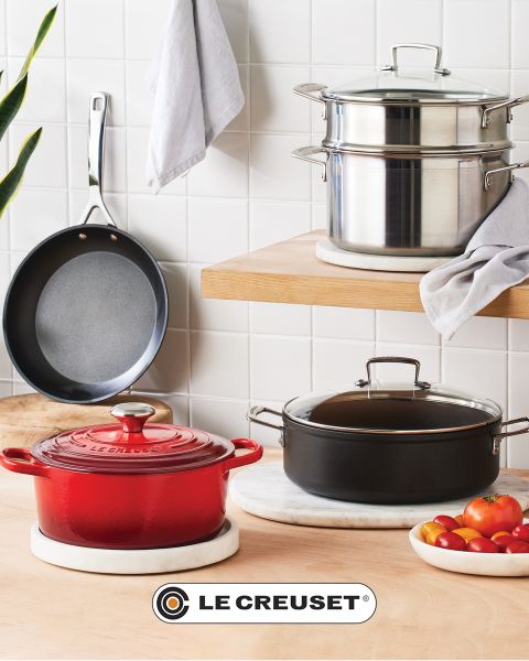 Eine Küchenszene mit einem roten Schmortopf, einer antihaftbeschichteten Bratpfanne, einem stapelbaren Topfset aus Edelstahl, einem schwarzen Kochtopf und einer Schüssel mit Kirschtomaten auf einer hellen Holztheke vor einer weißen Fliesenrückwand. Das Le Creuset-Logo ist sichtbar.