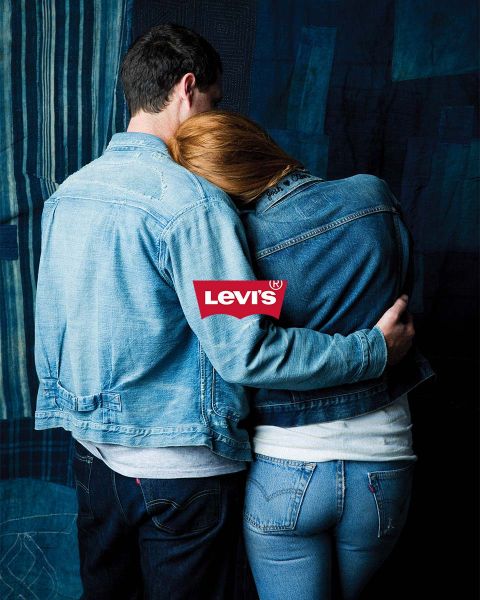Mann und Frau umarmen sich vor blauem Patchwork-Hintergrund, beide tragen Jeansjacken und Jeans. Über ihnen prangt deutlich ein rotes Levi's-Logo.