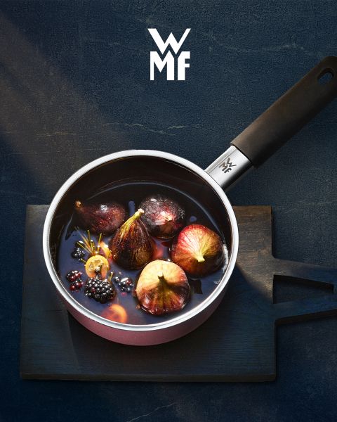 Ein mit Feigen, Brombeeren und aromatischen Garnituren gefüllter Kochtopf steht auf einem dunklen Holzbrett. Über dem Kochtopf ist das WMF-Logo zu sehen. Der Hintergrund ist eine strukturierte dunkle Oberfläche.