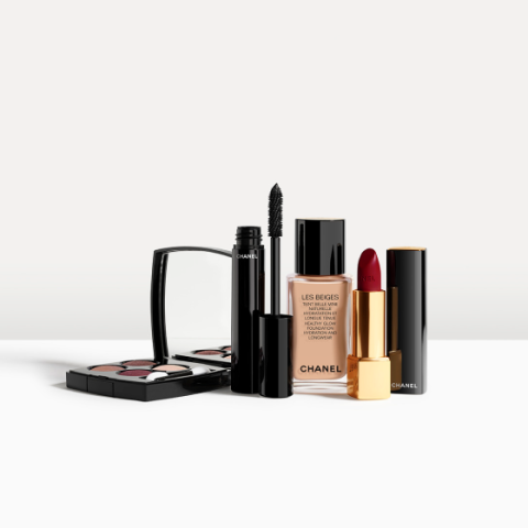 Eine Kollektion von Make-up-Produkten von Chanel, darunter ein Kompaktset mit Lidschatten, eine Wimperntusche, flüssige Grundierung in einer Glasflasche und ein dunkelroter Lippenstift in einer gold-schwarzen Hülle, angeordnet auf einer weißen Oberfläche.