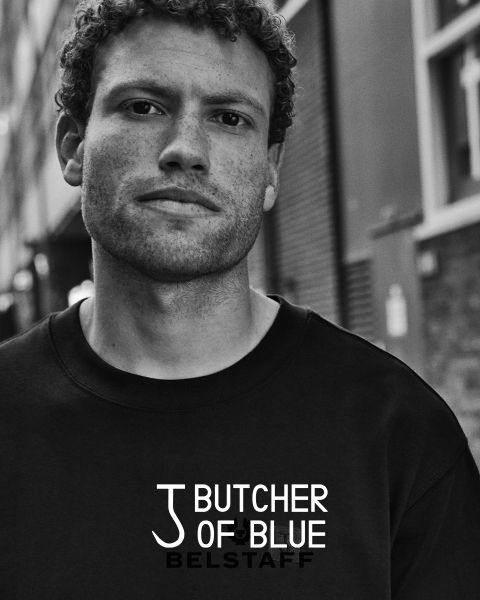 Schwarz-weißes Porträt eines Mannes mit kurzen lockigen Haaren, der ein Sweatshirt mit den Worten „Butcher of Blue“ und „Belstaff“ trägt. Er steht in einer städtischen Umgebung mit Gebäuden im Hintergrund.