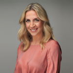 Eine Person mit langen, welligen blonden Haaren und einem warmen Lächeln trägt ein langärmliges, korallenfarbenes Oberteil. Der Hintergrund ist in einem neutralen Grau gehalten, was dem Bild eine professionelle und freundliche Ausstrahlung verleiht.