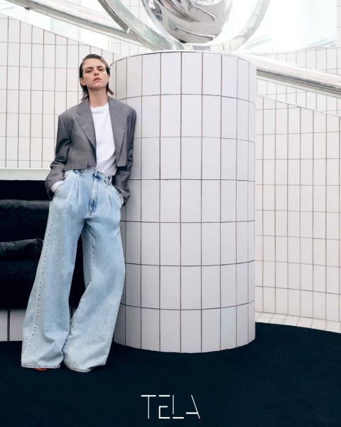 Person, die an einer weiß gefliesten Säule lehnt und einen grauen Blazer, ein weißes T-Shirt und übergroße hellblaue Jeans trägt. Der Boden ist schwarz und die Umgebung ist minimalistisch mit modernem Dekor. Unten steht das Wort „TELA“.