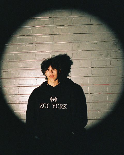 Eine Person mit lockigem Haar steht vor einer Backsteinmauer, die teilweise von einem kreisförmigen Licht beleuchtet wird. Sie trägt einen dunklen Kapuzenpullover mit dem Text „ZOO YORK“. Der Hintergrund liegt im Schatten, wodurch ein Spotlight-Effekt entsteht.